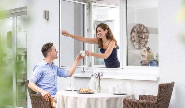 Schlanke Frau mit dunklen langen Haaren reicht eine Tasse Kaffee durch das geöffnete Fliegengitter Pendelfenster zu einem Mann der ausser vor dem Fenster am Tisch sitzt