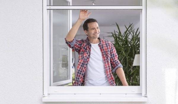 Mann mit dunklen Haaren, weissem Shirt und bunten Hemd steht am offenen Fenster und schiebt eine Fliegengitterrollo nach oben.