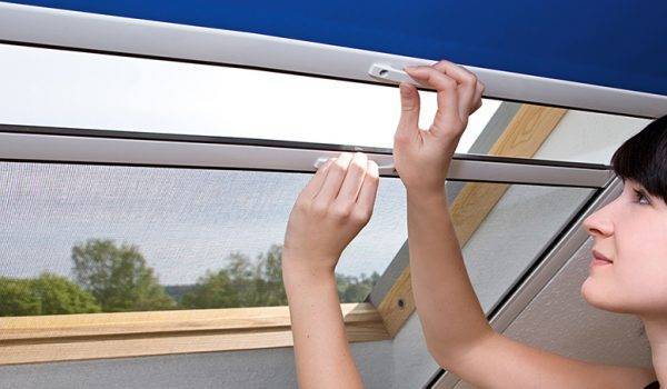 Eine junge Frau mit dunklen Haaren steht innen vor dem Dachfenster und hält mit der rechten Hand das obere blaue Sonnenschutzrollo und mit der linken das untere Fliegengitterrollo fest, um es zu verschieben.