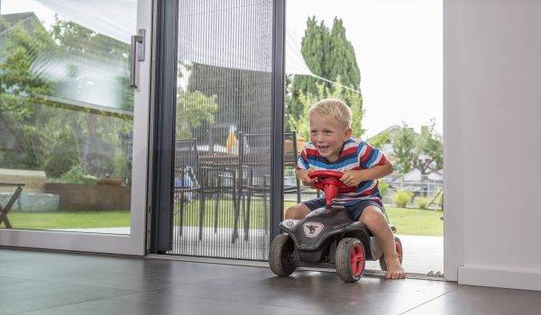 blonder Junge im Alter von 3 Jahren fährt mit seinem Bobbycar durch eine geöffnete Insektenschutz Plisseetür