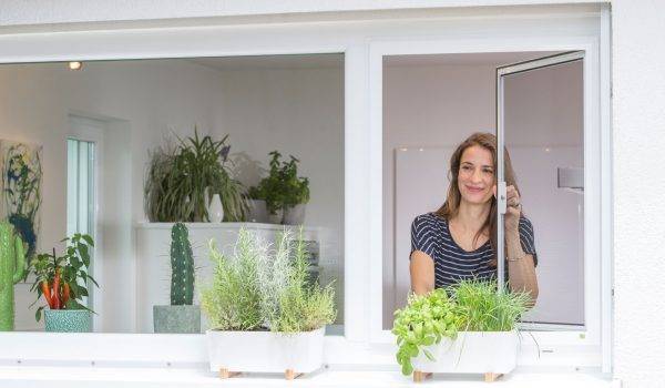 Junge Frau mit langen braunen Haaren öffnet ein Fenster mit Insektenschutzgitter und greift nach ihren Kräutern, welche aussen auf dem Fensterbrett in einer Blumenkiste wachsen