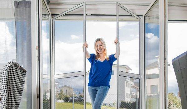 Sara Bachmann steht in einer halb geöffneten Fliegengitter Pendeltür mit 2 Flügeln und hält die Flügel in der Mitte fest