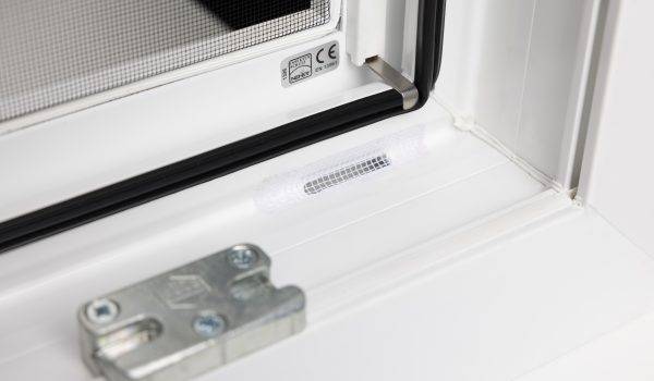 Ansicht eines weissen Kunststofffensters von innen. Man sieht einen Schlitz im Fensterprofil, welcher als Entwässerung dient. Darüber ist ein weisses Fliegengitterpflaster geklebt, um das Eindringen von Insekten zu verhindern.